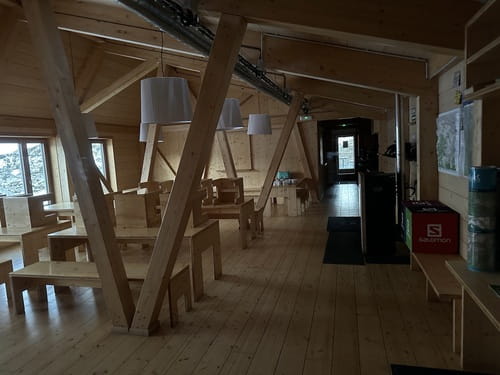 Intérieur en bois du refuge de l'Illa en Andorre, avec des tables en bois dans la salle à manger spacieuse à l'entrée. Un poêle réchauffe l'espace, tandis que des baies vitrées offrent une vue imprenable sur les sommets environnants.