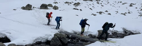 Quatre skieurs de randonnée traversant un pont sur un ruisseau dans les Encantats, Parc National d'Aigüestortes i Estany de Sant Maurici, entre neige et rochers, guidés par un professionnel de Sensations Pyrénées.