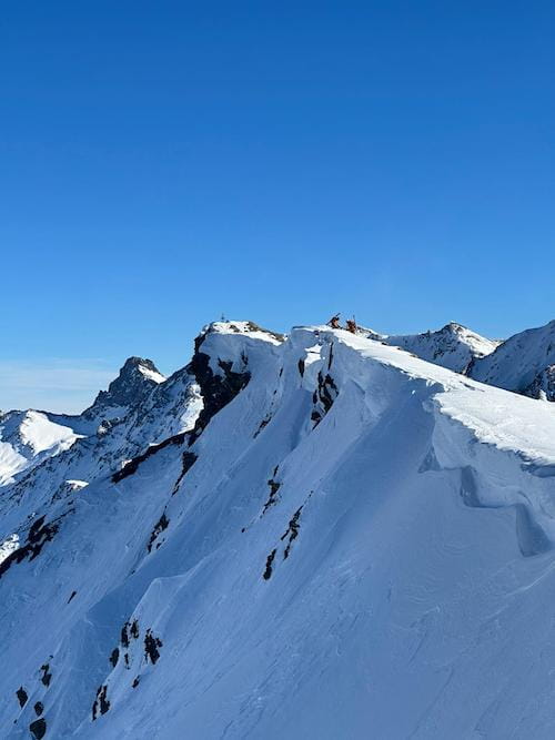 Deux alpinistes avec skis de randonnée sur le dos franchissent une crête pour atteindre un sommet dans le parc naturel du Queyras lors d'un raid à ski, illustrant l'aventure alpine accompagné d'un guide de haute montagne.