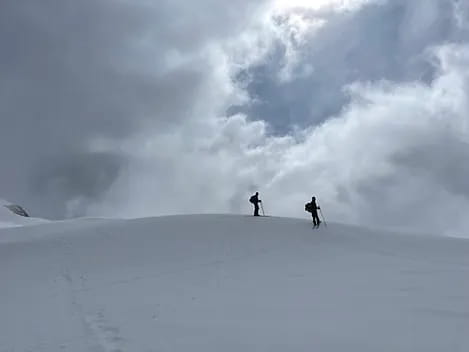 Paysage hivernal blanc et nuageux avec une éclaircie laissant passer les rayons du soleil, deux skieurs de randonnée redescendant le pic Aneto après l'avoir gravi.