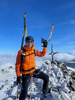 Skieur de randonnée flirtant avec l'alpinisme, souriant et fier avec baudrier encordé et piolet, au sommet du Grand Péric, croix enneigée en arrière-plan.